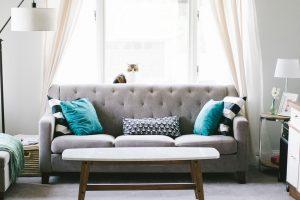 A close up of a grey couch with blue pillows. There is a coffee table in front of it and a window with white curtains behind it. 