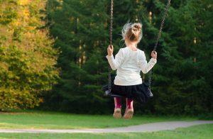 A girl in a white shirt, black skirt and pink rain-boots on a swing in some natural setting. 