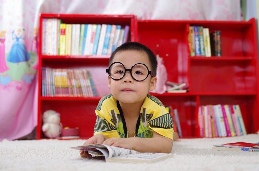 A boy with a book in front of him, and red bookshelf behind him. Moving during the school year is particularly hard for young kids.