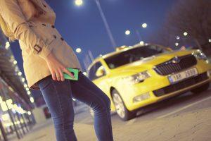 A woman holding a green phone and a yellow taxi in the background, heading in her direction. 