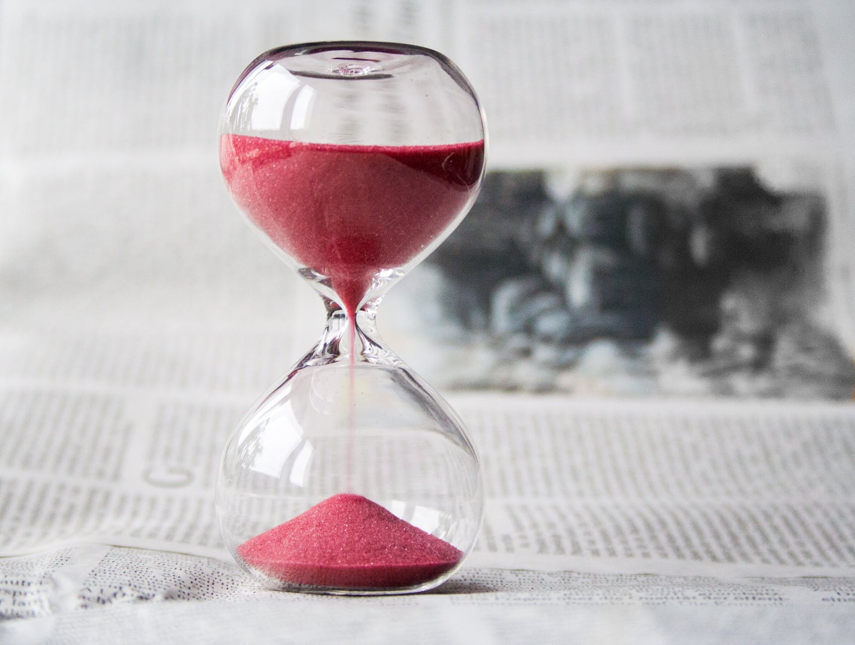An hourglass with red sand inside. It is placed on top of some surface which seems to be covered in newspapers. When you have to pack in 7 days, every second counts.
