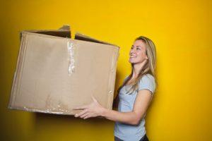 A woman holding a big cardboard box and standing in front of a yellow wall. She is smiling and wearing a grey t-shirt.