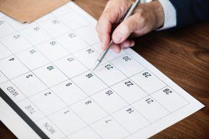 A calendar and a person`s hand holding a pen. He might be marking a certain date in the calendar, and panicking that he has to pack in 7 days.